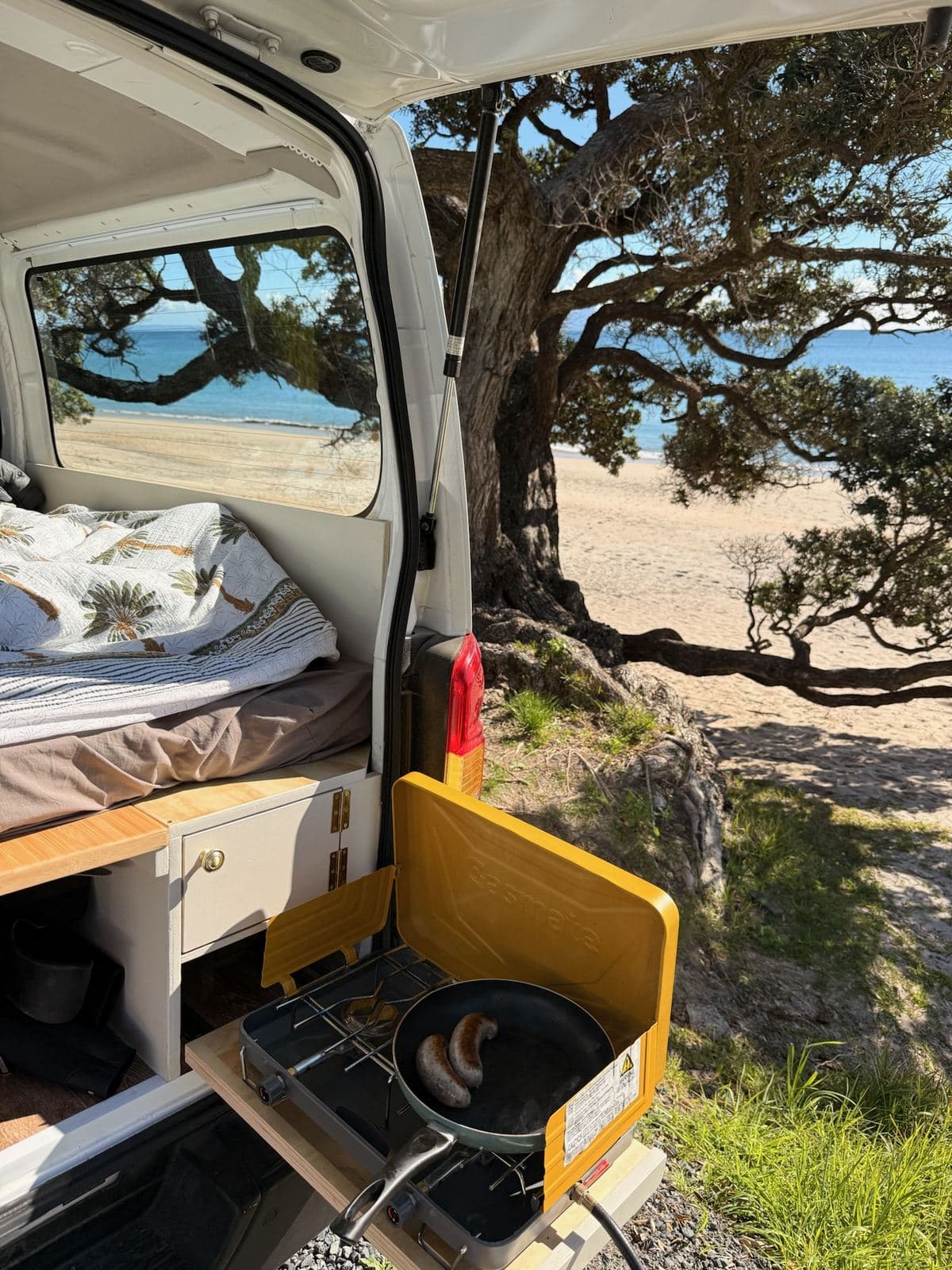 The Pīwakawaka's pull-out kitchen with a gas hob in use at the rear, beach and pōhutukawa beyond