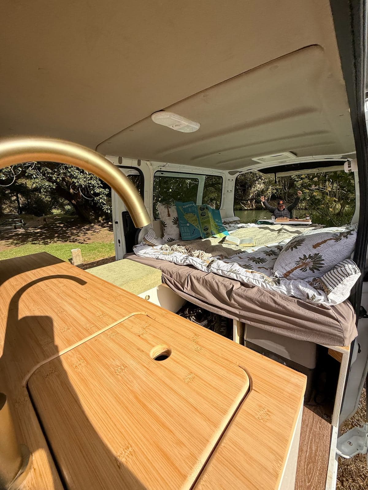 The Pīwakawaka's worktop and sink lid with the bed made up behind and the side and rear doors open to a campsite