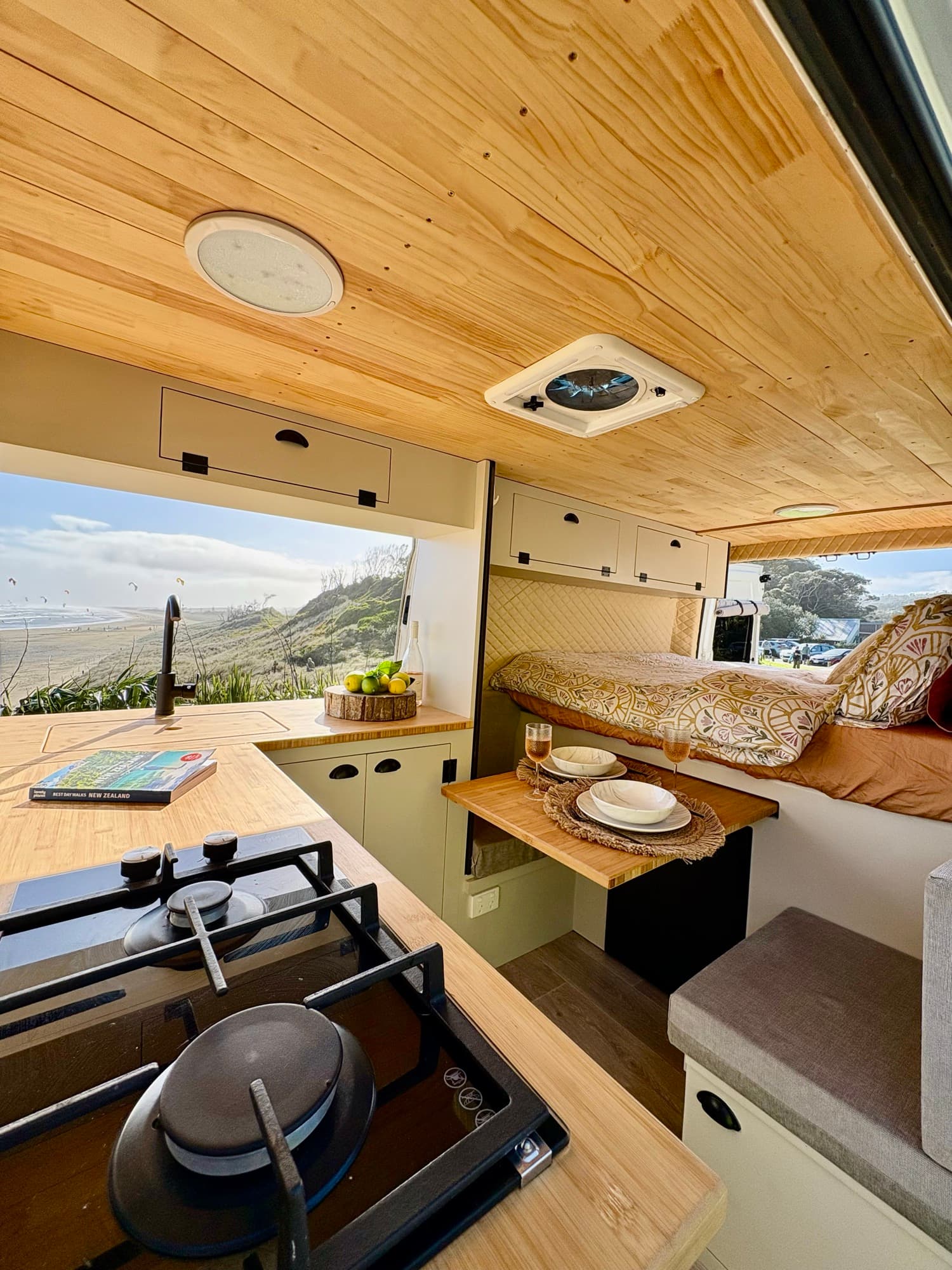 Inside the Tūī 2-berth campervan — hob, kitchen, dinette and double bed with a sea view