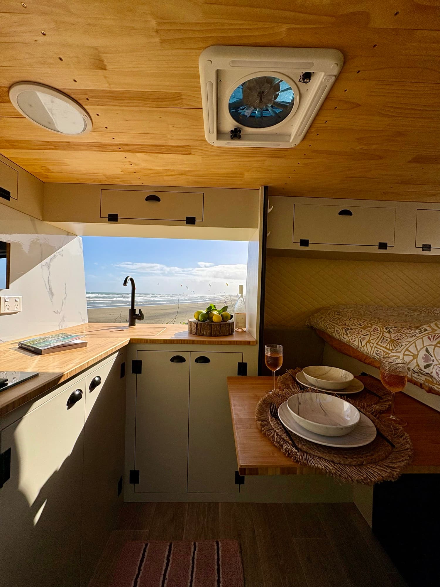 Tūī 2-berth campervan galley kitchen and double bed with a beach view through the window