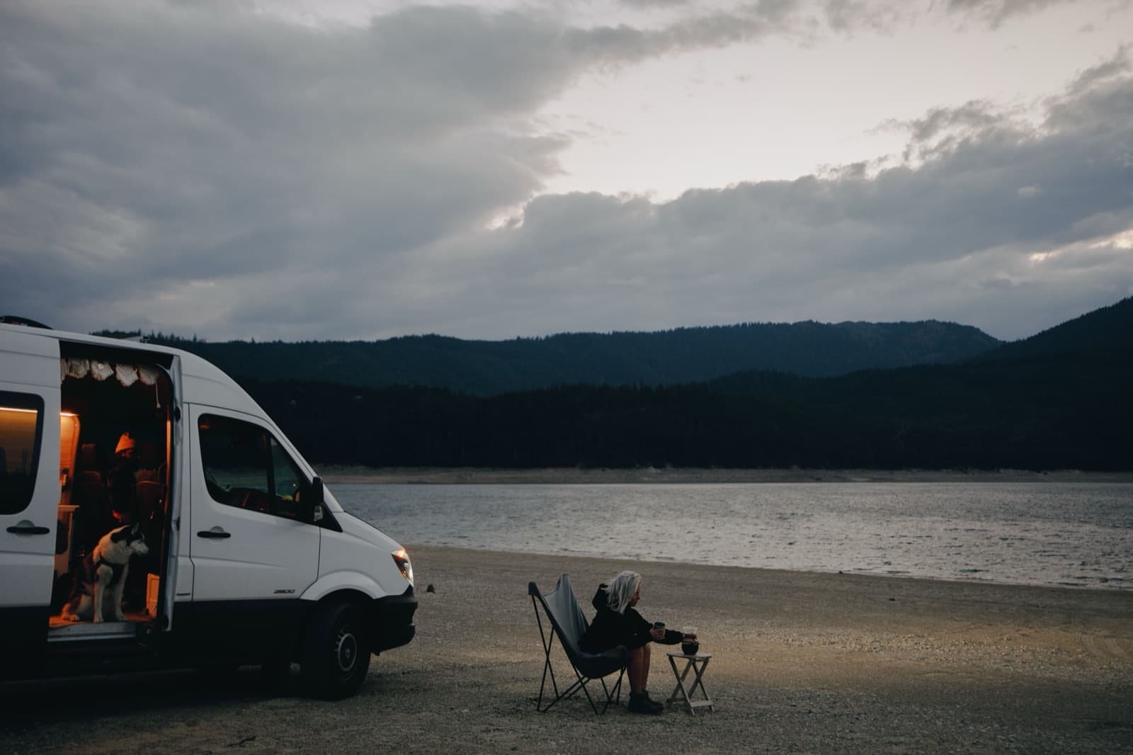 Person sitting beside an unmarked campervan at a lake, freedom camping in New Zealand